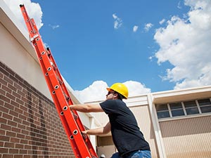 Construction worker wearing a yellow hard hat climbs a red ladder against a brick wall under a blue sky.