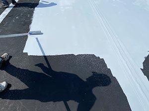 Roof-Restoration-Bowling-Green-KY-Kentucky-1 | Glick Roofing Systems Person applying light blue paint to a dark roof with a roller; shadow of the painter visible on the surface.