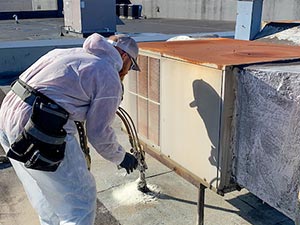 Spray-Foam-Roofing-Bowling-Green-KY-Kentucky-1 | Glick Roofing Systems Worker in a white protective suit and cap applies sealant along a rooftop edge with a caulking gun near an air conditioning unit.