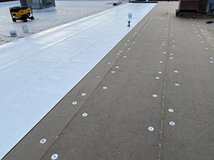Roof being installed: white underlayment sheet laid to the left, dark roof deck on the right with many round fasteners visible.